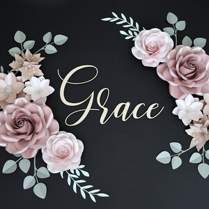 May include: A paper flower arrangement with the name "Grace" in the center. The flowers are in shades of pink and white, with green leaves. The flowers are arranged in a semi-circle shape.