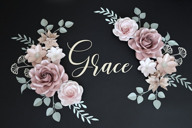 May include: A paper flower arrangement with the name "Grace" in the center. The flowers are in shades of pink and white, with green leaves. The flowers are arranged in a semi-circle shape.