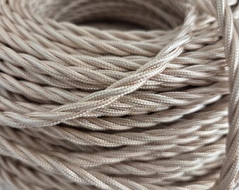 May include: A spool of beige fabric-covered electrical wire featuring a twisted, braided design. The cord has a smooth, slightly reflective texture and is coiled tightly in a circular formation, showing the intricate weave of the textile casing.