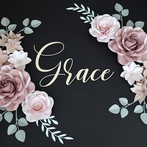 May include: A paper flower arrangement with the word "Grace" in the center. The flowers are in shades of pink and white, with green leaves. The flowers are arranged in a circular pattern around the word "Grace".