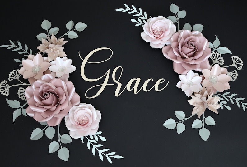 May include: A paper flower arrangement with the word "Grace" in the center. The flowers are in shades of pink and white, with green leaves. The flowers are arranged in a circular pattern around the word "Grace".