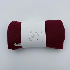 May include: A rolled-up burgundy fabric item with a white label wrapped around it. The label reads "curlyncovered" and "MADE IN GERMANY" with a leaf illustration. The website address www.curlyncovered.com is also visible.