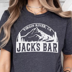 May include: Dark grey t-shirt featuring the text "VIRGIN RIVER, CA" above a mountain graphic and the words "JACKS BAR" in a white, arched design. The shirt is short-sleeved and made of a soft material.