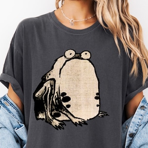 Puede incluir: Una camiseta gris oscuro con una ilustraci&oacute;n en blanco y negro de una rana con ojos grandes.