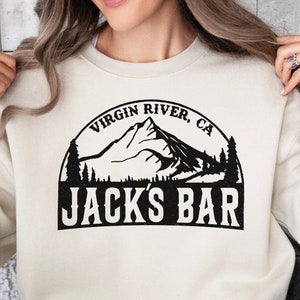May include: A cream-coloured sweatshirt with a black graphic design of a mountain range with trees and the text "Virgin River, CA Jack's Bar".