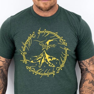 May include: A dark green t-shirt with a yellow graphic of a mountain, a flying creature, and the inscription "One Ring to rule them all, One Ring to find them, One Ring to bring them all and in the darkness bind them."