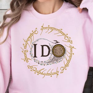 May include: A light pink sweatshirt with a gold circular design featuring the text "IDO" and a hobbit door illustration. The design is surrounded by script. The sweatshirt is a long-sleeved crew neck.