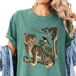 May include: A teal-colored t-shirt featuring a vintage-style illustration of a tiger with orange eyes and black stripes. The tiger is depicted with a bird perched on a tree branch. The shirt has short sleeves and a relaxed fit.