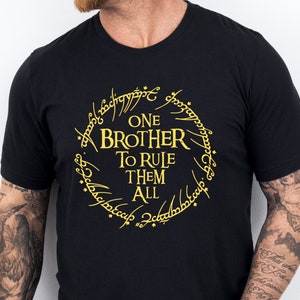 Op de afbeelding: Zwart T-shirt met een gouden afbeelding van de Ene Ring uit De Heer der Ringen. De afbeelding zegt "One Brother To Rule Them All".
