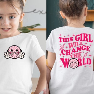 May include: White t-shirt with a pink and white checkered smiley face wearing sunglasses and a peace sign. The back of the shirt has the text "THIS GIRL WILL CHANGE THE WORLD" in pink.