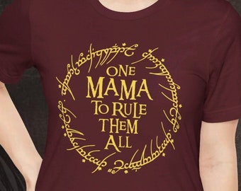 May include: Burgundy t-shirt with a gold ring design and the text "ONE MAMA TO RULE THEM ALL". The design is a circular pattern with stylized text. The shirt has short sleeves and a crew neck.