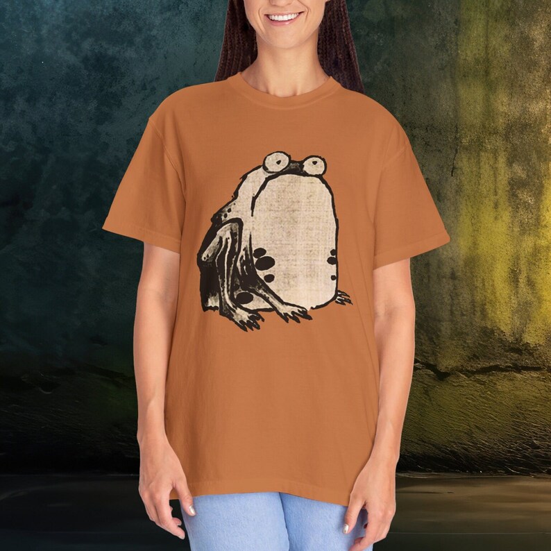 Puede incluir: Una camiseta marr&oacute;n con una ilustraci&oacute;n en blanco y negro de una rana en la parte delantera.