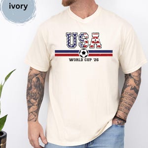 May include: An ivory short-sleeve t-shirt featuring a graphic print with the text USA in a distressed American flag pattern, a horizontal blue and red stripe, a soccer ball icon, and the text WORLD CUP '26 centered on the chest.