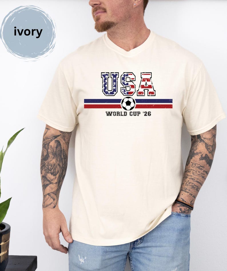 May include: An ivory short-sleeve t-shirt featuring a graphic print with the text USA in a distressed American flag pattern, a horizontal blue and red stripe, a soccer ball icon, and the text WORLD CUP '26 centered on the chest.