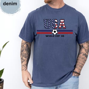 May include: A denim blue short-sleeve t-shirt featuring a graphic with the text USA in a red, white, and blue flag pattern, a soccer ball, and the text WORLD CUP '26 centered above two horizontal stripes in dark blue and red colors.
