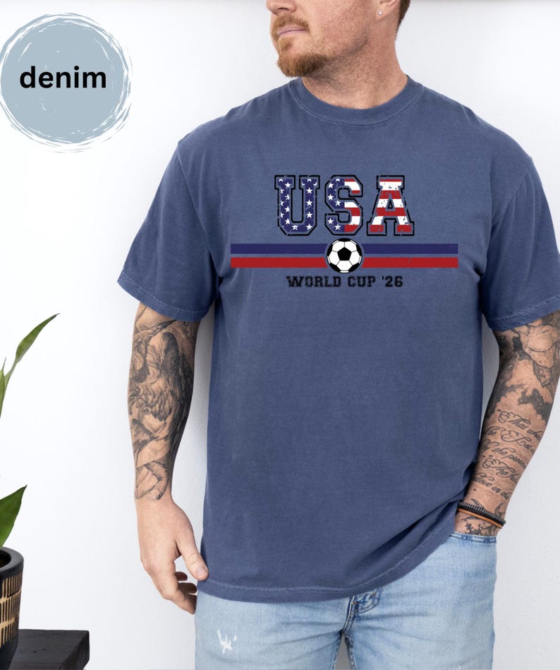 May include: A denim blue short-sleeve t-shirt featuring a graphic with the text USA in a red, white, and blue flag pattern, a soccer ball, and the text WORLD CUP '26 centered above two horizontal stripes in dark blue and red colors.