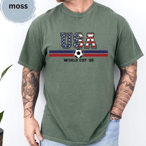 May include: A moss green short-sleeve t-shirt featuring the text USA in a distressed American flag pattern, a soccer ball graphic, and the text WORLD CUP '26 centered above two horizontal stripes in dark blue and red across the chest area.