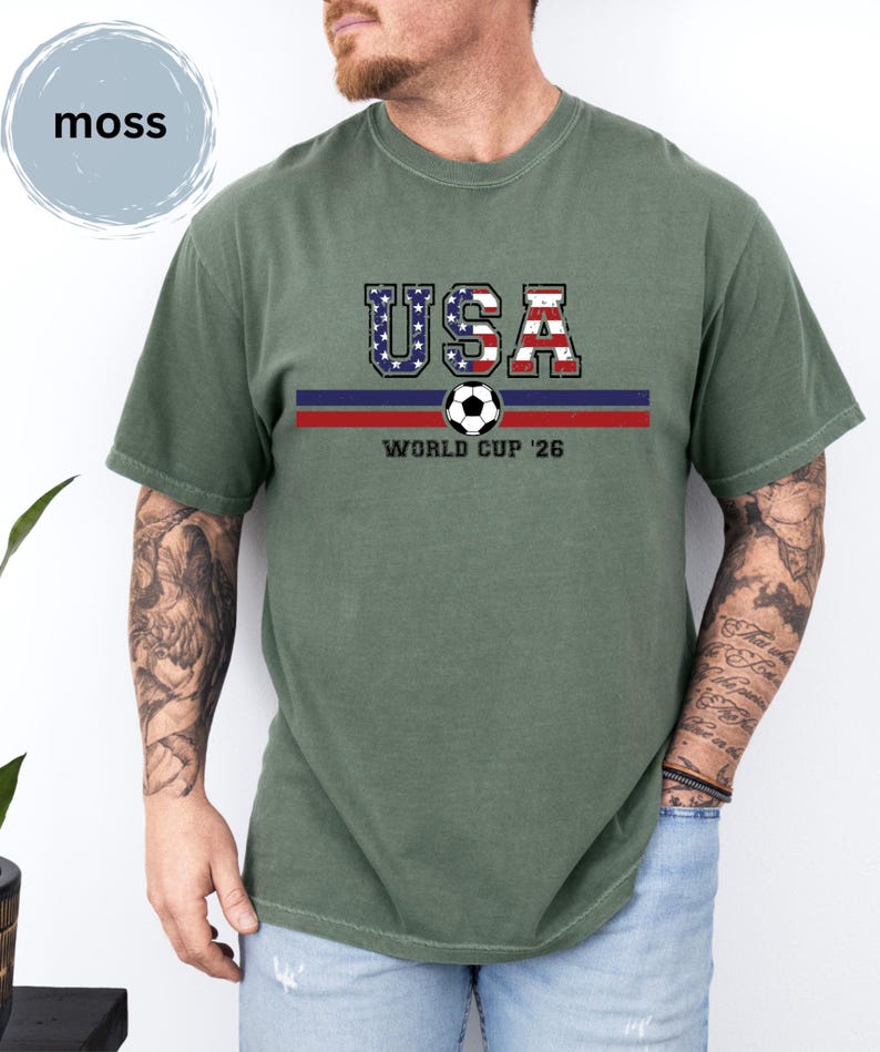 May include: A moss green short-sleeve t-shirt featuring the text USA in a distressed American flag pattern, a soccer ball graphic, and the text WORLD CUP '26 centered above two horizontal stripes in dark blue and red across the chest area.