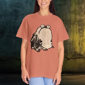 Puede incluir: Una camiseta de color coral con una ilustraci&oacute;n en blanco y negro de una rana en la parte delantera.