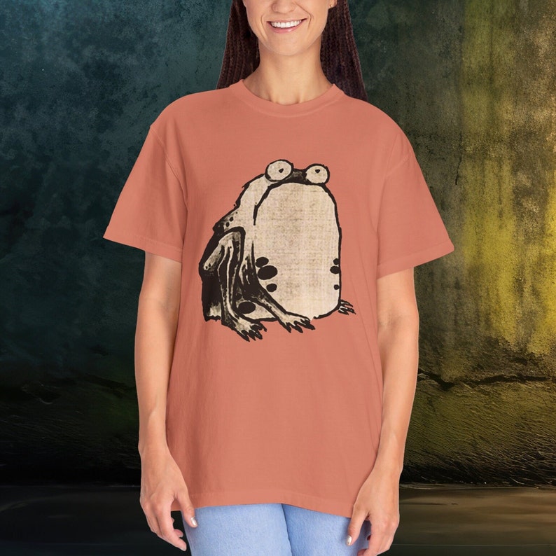 Puede incluir: Una camiseta de color coral con una ilustraci&oacute;n en blanco y negro de una rana en la parte delantera.