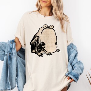 Puede incluir: Una camiseta de color crema con una ilustraci&oacute;n en blanco y negro de una rana con ojos grandes.