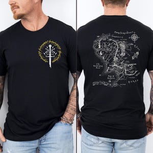 Pode incluir: T-shirt preto com um design dourado e branco na frente, incluindo uma espada e um emblema circular com texto. A parte de trás da camisa exibe um mapa detalhado com vários locais e texto. A camisa é feita de um material macio.
