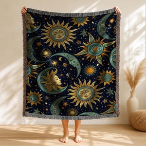 May include: A dark blue throw blanket featuring a celestial design with smiling suns, crescent moons, and stars in shades of gold, teal, and white. The blanket has a fringed edge and is held up by a person.