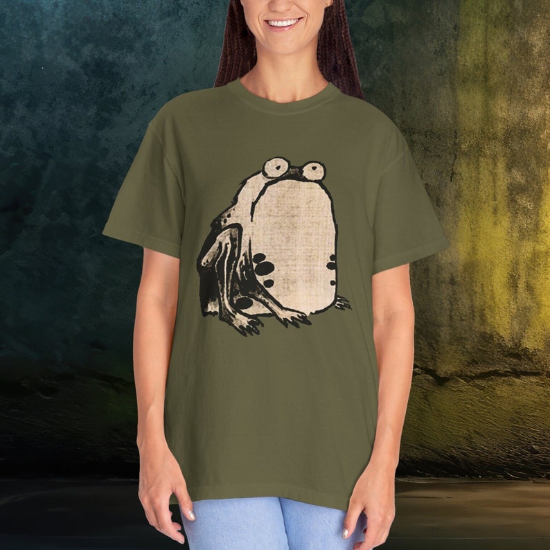 Puede incluir: Una camiseta verde con una ilustraci&oacute;n en blanco y negro de una rana con ojos grandes.