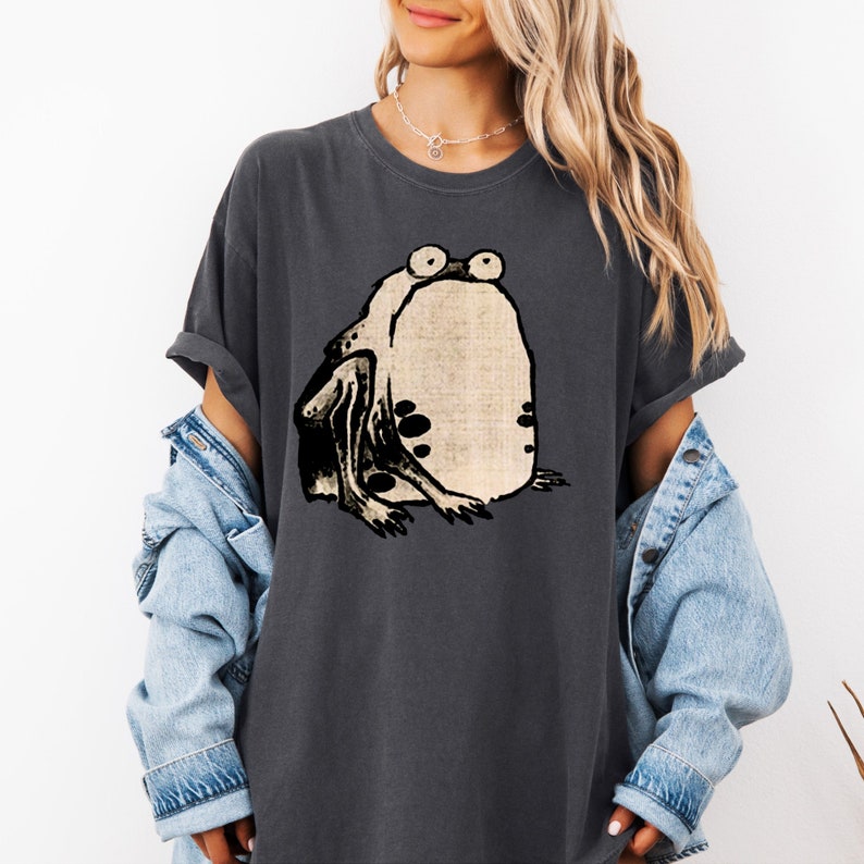 Puede incluir: Una camiseta gris oscuro con una ilustraci&oacute;n en blanco y negro de una rana con ojos grandes.