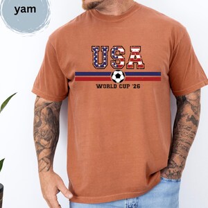 May include: A rust-colored short-sleeve t-shirt featuring the text USA in a distressed American flag pattern above a horizontal blue and red stripe. A soccer ball graphic is centered on the stripe with the text WORLD CUP '26 printed below it.
