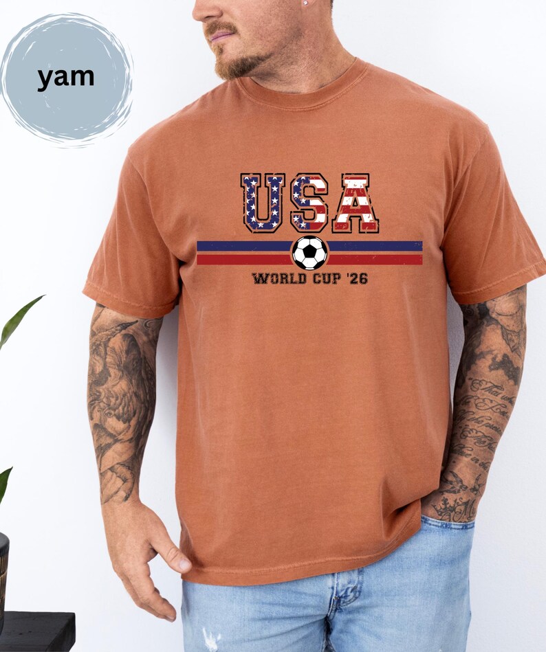 May include: A rust-colored short-sleeve t-shirt featuring the text USA in a distressed American flag pattern above a horizontal blue and red stripe. A soccer ball graphic is centered on the stripe with the text WORLD CUP '26 printed below it.