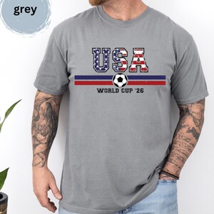 May include: A grey short-sleeved t-shirt featuring the text USA in a distressed American flag pattern, a horizontal blue and red stripe, a soccer ball graphic, and the text WORLD CUP '26 printed in black on the front of the garment.