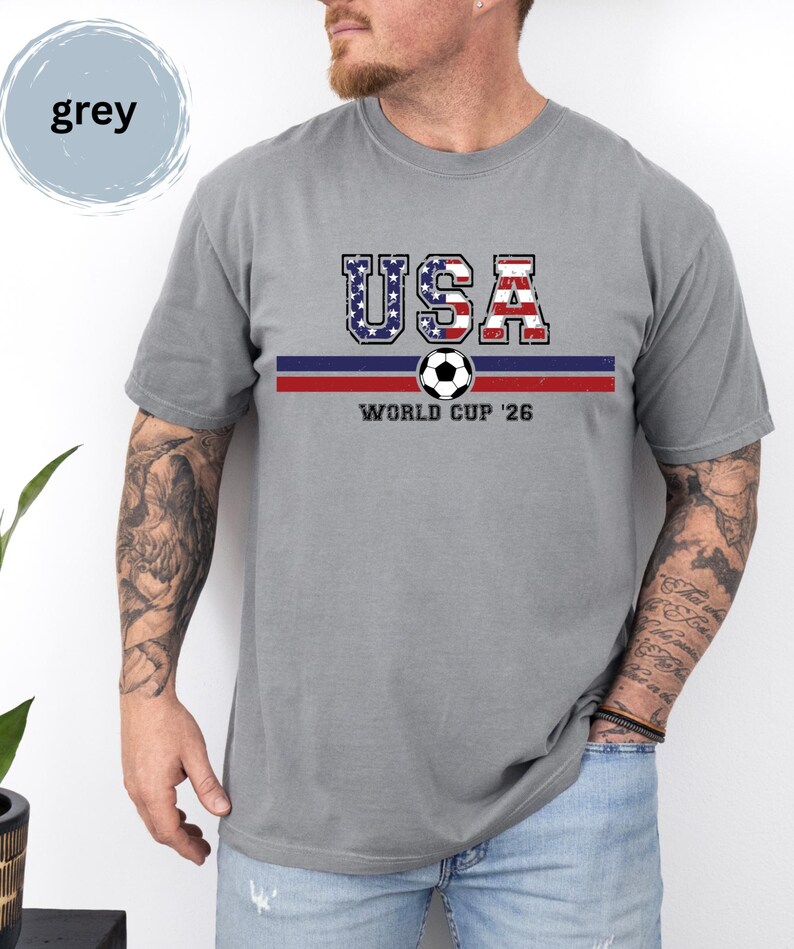 May include: A grey short-sleeved t-shirt featuring the text USA in a distressed American flag pattern, a horizontal blue and red stripe, a soccer ball graphic, and the text WORLD CUP '26 printed in black on the front of the garment.