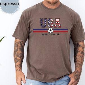 May include: An espresso-colored short-sleeve t-shirt featuring a graphic print with the text USA in a distressed American flag pattern, a soccer ball icon, horizontal blue and red stripes, and the text WORLD CUP '26 centered on the chest.
