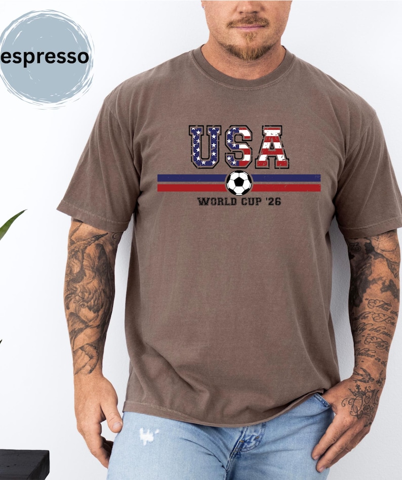 May include: An espresso-colored short-sleeve t-shirt featuring a graphic print with the text USA in a distressed American flag pattern, a soccer ball icon, horizontal blue and red stripes, and the text WORLD CUP '26 centered on the chest.
