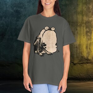 Puede incluir: Una camiseta gris antracita con una ilustraci&oacute;n vintage de una rana con ojos grandes. La rana est&aacute; sentada sobre sus patas traseras con las patas delanteras cruzadas.