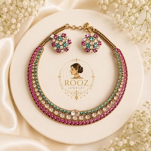 May include: A gold-toned necklace and matching earrings set with pink, teal, and clear gemstones. The necklace and earrings are arranged on a white plate with the "ROOZ JEWELRY" logo. White flowers are in the background.