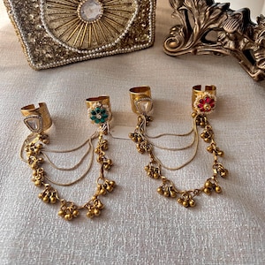 May include: Gold-toned finger rings with dangling chains and small bell charms. Each ring features a decorative top with a gemstone or floral design. The rings are displayed on a textured, light-colored surface.