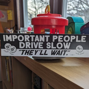 May include: Black and white bumper sticker with the text "IMPORTANT PEOPLE DRIVE SLOW" and "THEY'LL WAIT."  The sticker has a design of two 5-cent coins and a 10-cent coin.