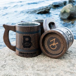 May include: Wooden beer mugs with stainless steel interiors. One mug features the letter "B" and the word "BROWNS" with "GROOMSMAN 10.20.2025" below. Another mug has the initials "ZD" and a Celtic knot design. The mugs are brown with black accents.