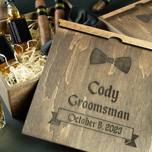 May include: A wooden box with a lid engraved with the text "Cody Groomsman October 5, 2023" and a bow tie design. The box contains two small bottles of liquor, cigars, and a bow tie.