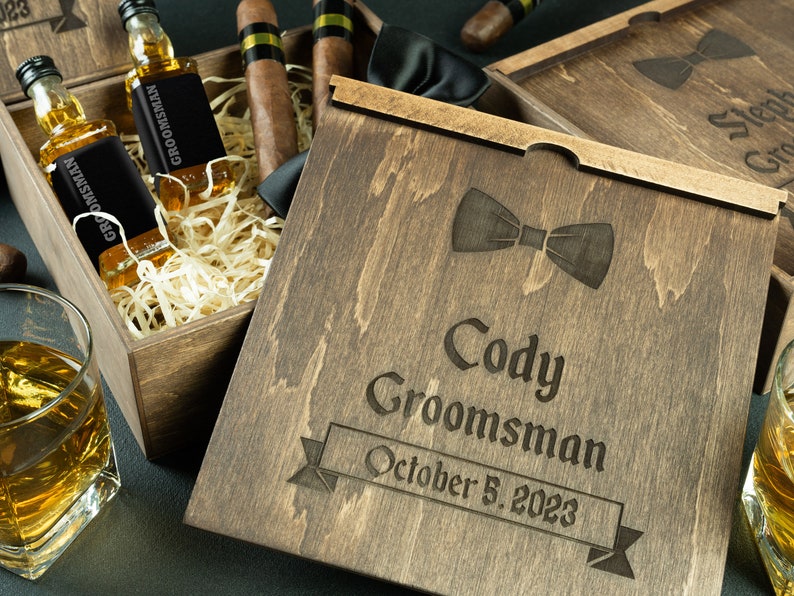 May include: A wooden box with a lid engraved with the text "Cody Groomsman October 5, 2023" and a bow tie design. The box contains two small bottles of liquor, cigars, and a bow tie.