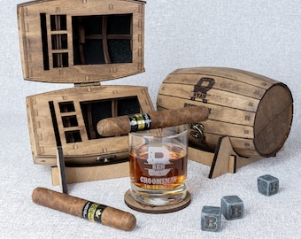 May include: A wooden barrel-shaped box with a hinged lid, a glass with the word "Groomsman" engraved on it, a cigar, and four gray stone ice cubes. The box is engraved with the name "Ryan" and the word "Whiskey".