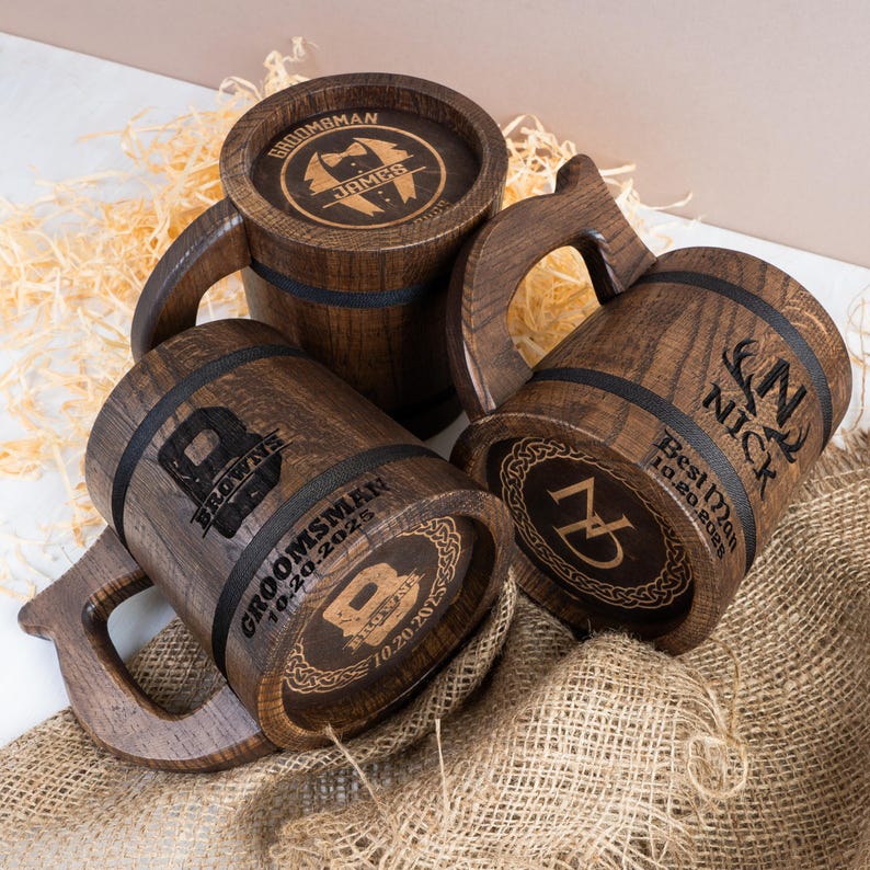 May include: Three wooden beer tankards with dark brown finishes and black bands. Each tankard has a handle and personalised engravings, including names and dates. The tankards are displayed on a burlap surface with decorative straw.
