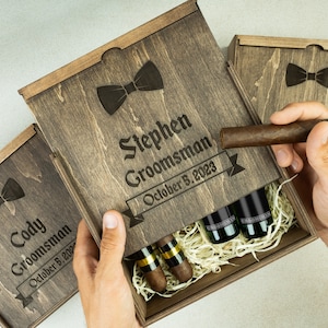 May include: Two wooden gift boxes with engraved bowties and the text "Cody Groomman October 8, 2023" and "Stephen Groomman October 8, 2023". The boxes are open and contain small bottles and cigars.
