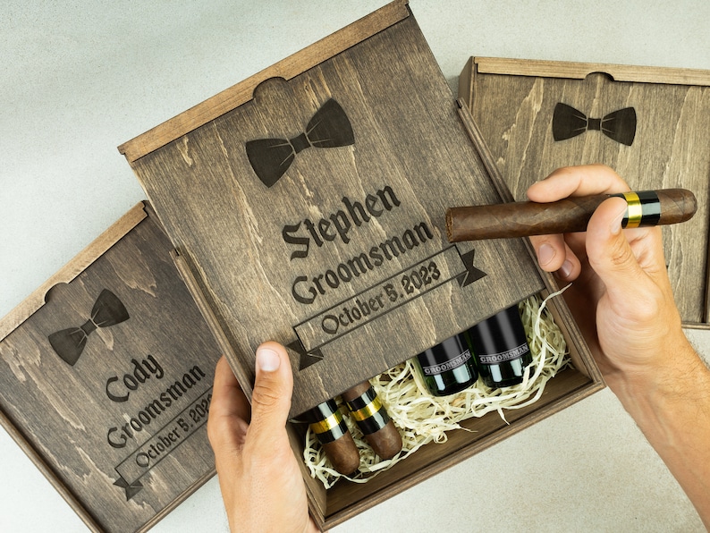 May include: Two wooden gift boxes with engraved bowties and the text "Cody Groomman October 8, 2023" and "Stephen Groomman October 8, 2023". The boxes are open and contain small bottles and cigars.