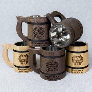 May include: Several wooden beer tankards with stainless steel interiors. Some are engraved with names, titles, and dates, including "Groomsman" and "Best Man." The mugs have dark brown and light wood tones with black accents.