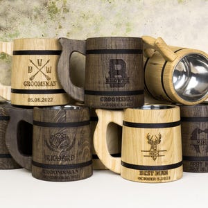 May include: A selection of wooden beer tankards with stainless steel interiors. The tankards are engraved with names, dates, and titles such as "Groomsman" and "Best Man." The mugs have a rustic, handcrafted appearance.
