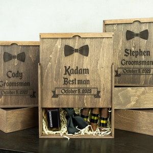 May include: Three wooden boxes with engraved names, dates, and bowties. The middle box is open and contains a black bowtie, a small bottle, and cigars.