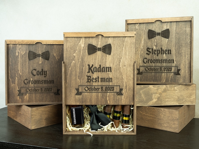 May include: Three wooden boxes with engraved names, dates, and bowties. The middle box is open and contains a black bowtie, a small bottle, and cigars.